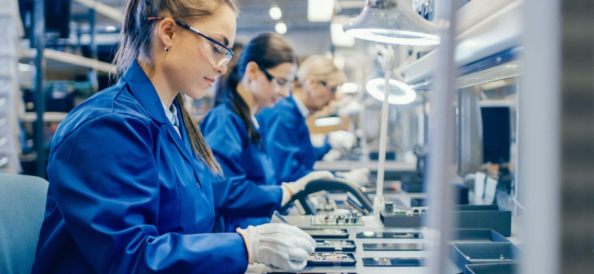 Female Electronics Factory Workers in Blue Work Coat and Protective Glasses Assembling Printed Circuit Boards for Smartphones with Tweezers. High Tech Factory with more Employees in the Background.