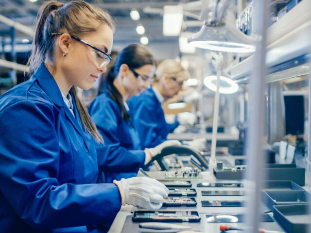 Female Electronics Factory Workers in Blue Work Coat and Protective Glasses Assembling Printed Circuit Boards for Smartphones with Tweezers. High Tech Factory with more Employees in the Background.